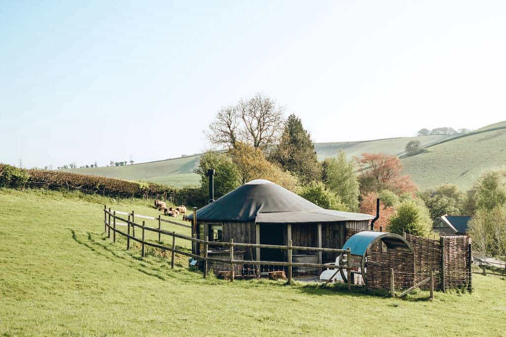 A circular timber Roundhouse with a black conical roof nestled in a green valley with sheep grazing nearby.