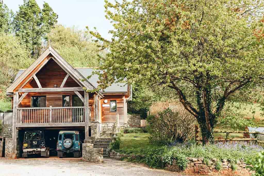 Exterior of the timber-clad cottage featuring a stone staircase and a car port with two vintage Land Rovers parked inside.